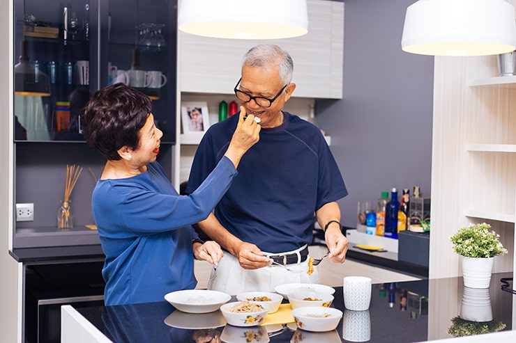 Senior Asian couple grandparents cooking together while woman is feeding food to man at the kitchen