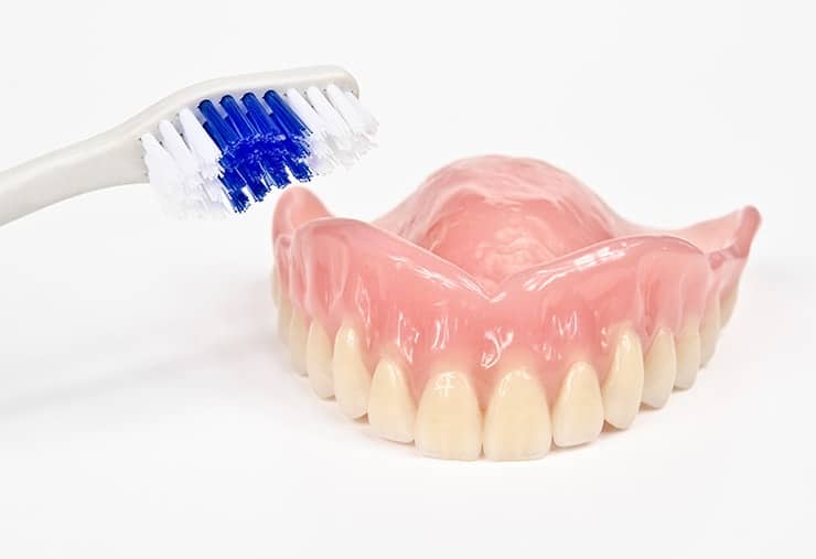 An upper denture sitting on a countertop with a toothbrush. Now that you have a new set of dentures it's time to have a daily care schedule.